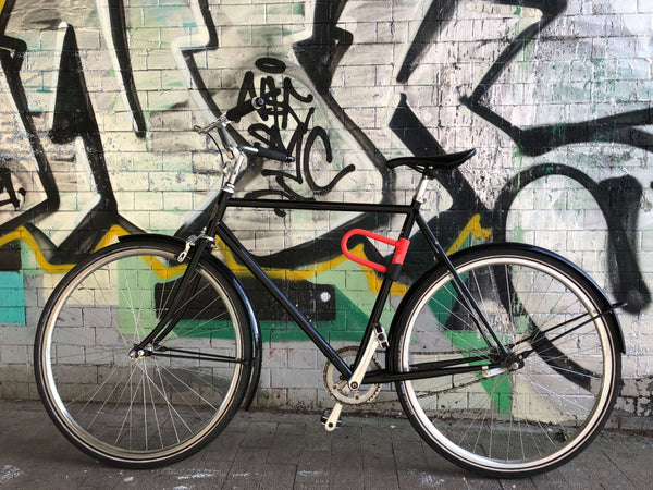 A black bicycle with a hardened steel casing leaning against a brick wall, secured with the CYCL Red Lock and U-Lock.